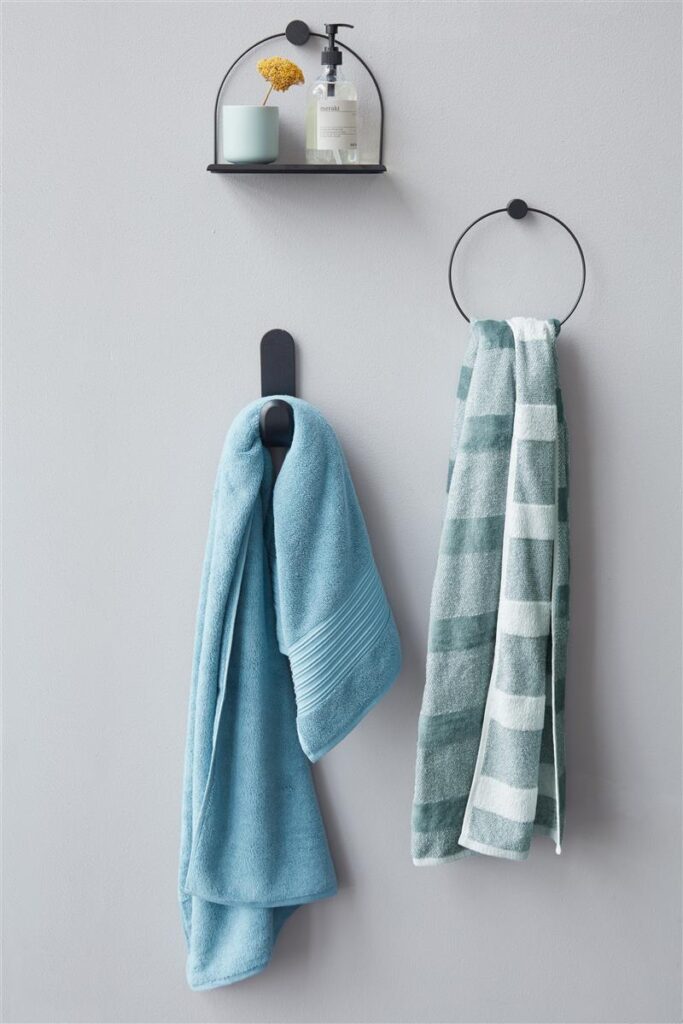 Twee Beddinghouse Sheer Gastendoek Blauw-handdoeken hangen aan een lichtgrijze muur onder een zwarte plank met accessoires.