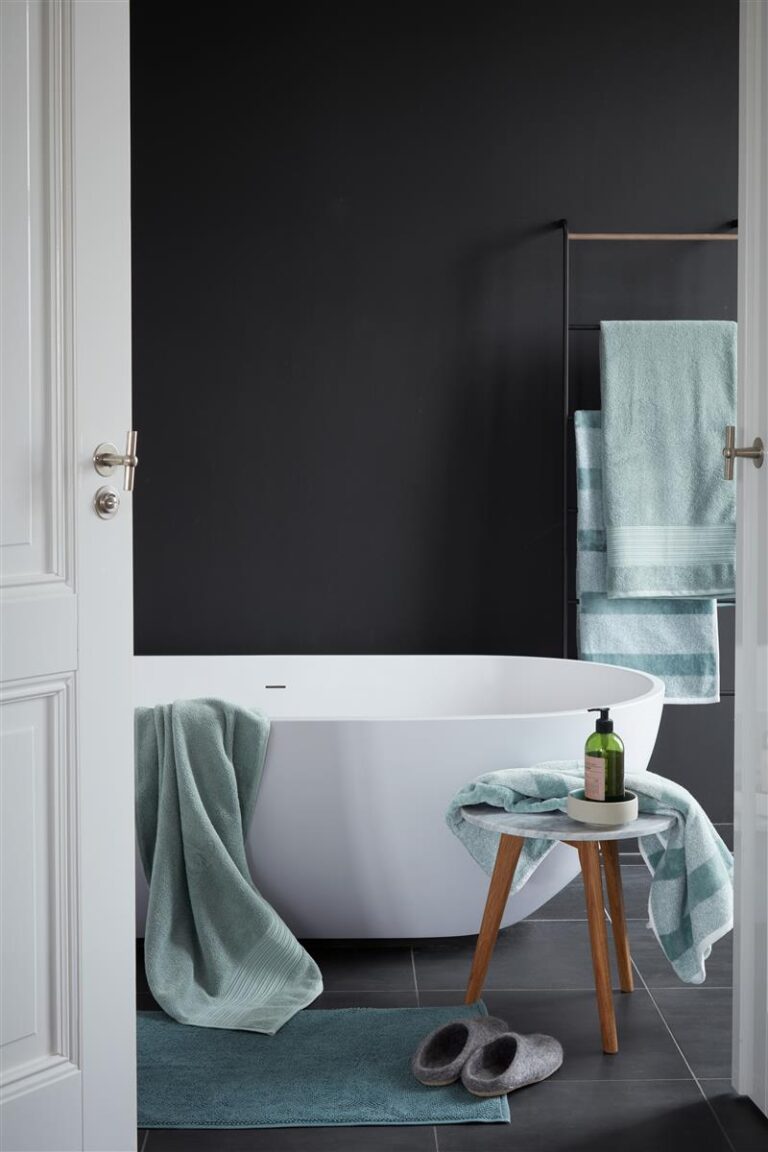 Een moderne badkamer met een vrijstaand bad, blauwgroene handdoeken, de Beddinghouse Sheer Washand Groen-set, grijze badslippers en een houten krukje.