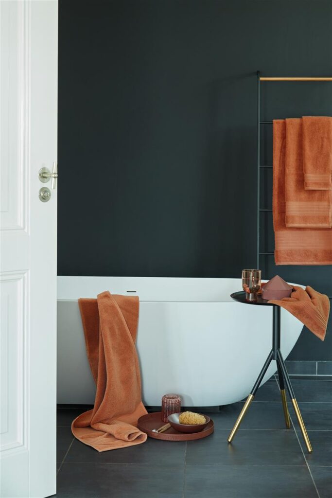 Een moderne badkamer met donkere muren heeft een wit bad, roestoranje Beddinghouse Sheer Gastendoek Oranje handdoeken op een rek en langs de rand, een zwart tafeltje met goudkleurige poten en een donkere tegelvloer.