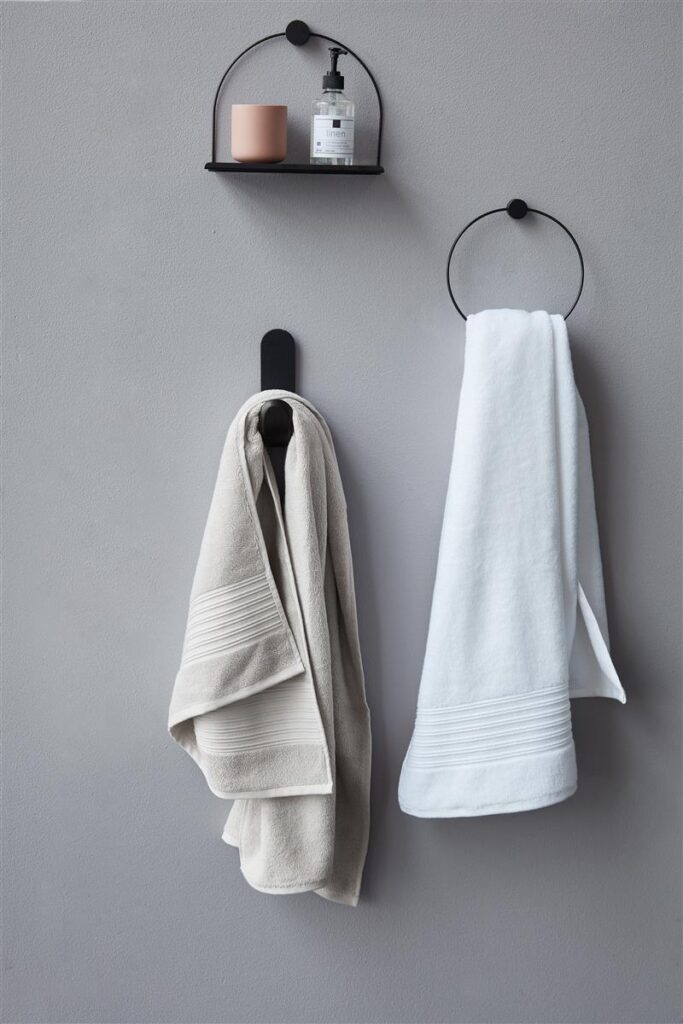 Het witte, doorschijnende badlaken van Beddinghouse hangt aan een grijze muur en zorgt voor een minimalistische uitstraling onder een zwart plankje met daarop een roze beker en een doorzichtige zeepdispenser.