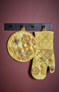 De gele ovenwant met Van Gogh-zonnebloemen hangt sierlijk aan een haak tegen een warme kastanjebruine muur.