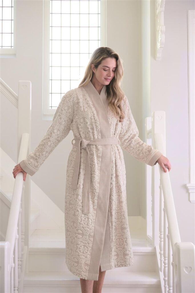 Een vrouw draagt de elegante Pip Studio Tile de Pip-badjas in kleur Khaki M en glimlacht zachtjes in een stijlvolle kamer met hoge ramen.