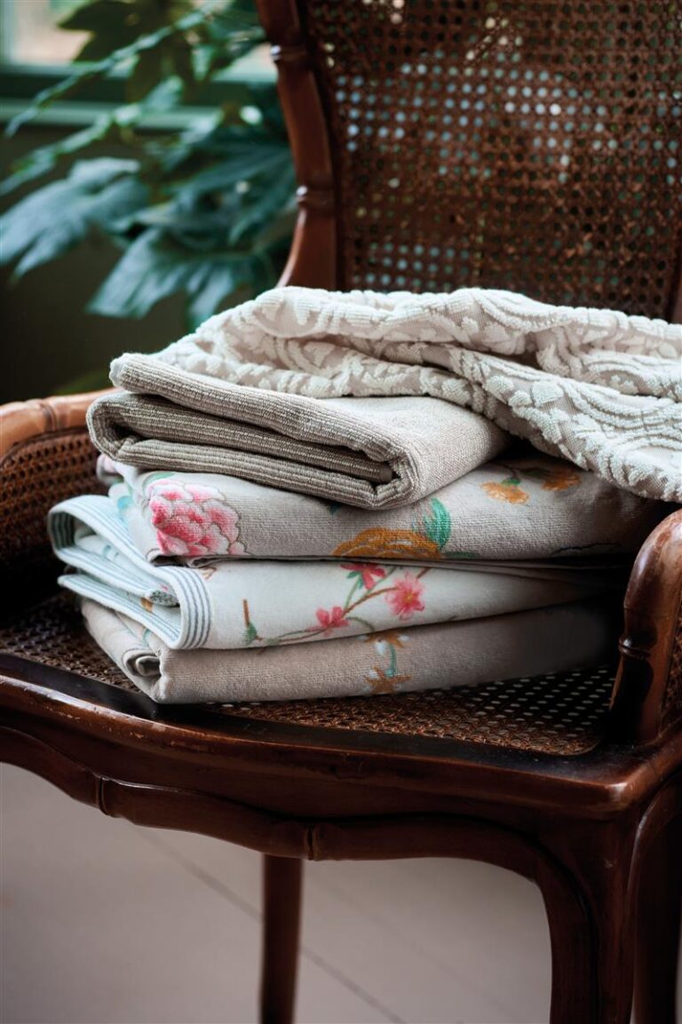 Op een bruine rotanstoel liggen drie netjes opgevouwen Pip Studio Tile de Pip Washand Khaki-handdoeken, met op de achtergrond vaag een groene plant.