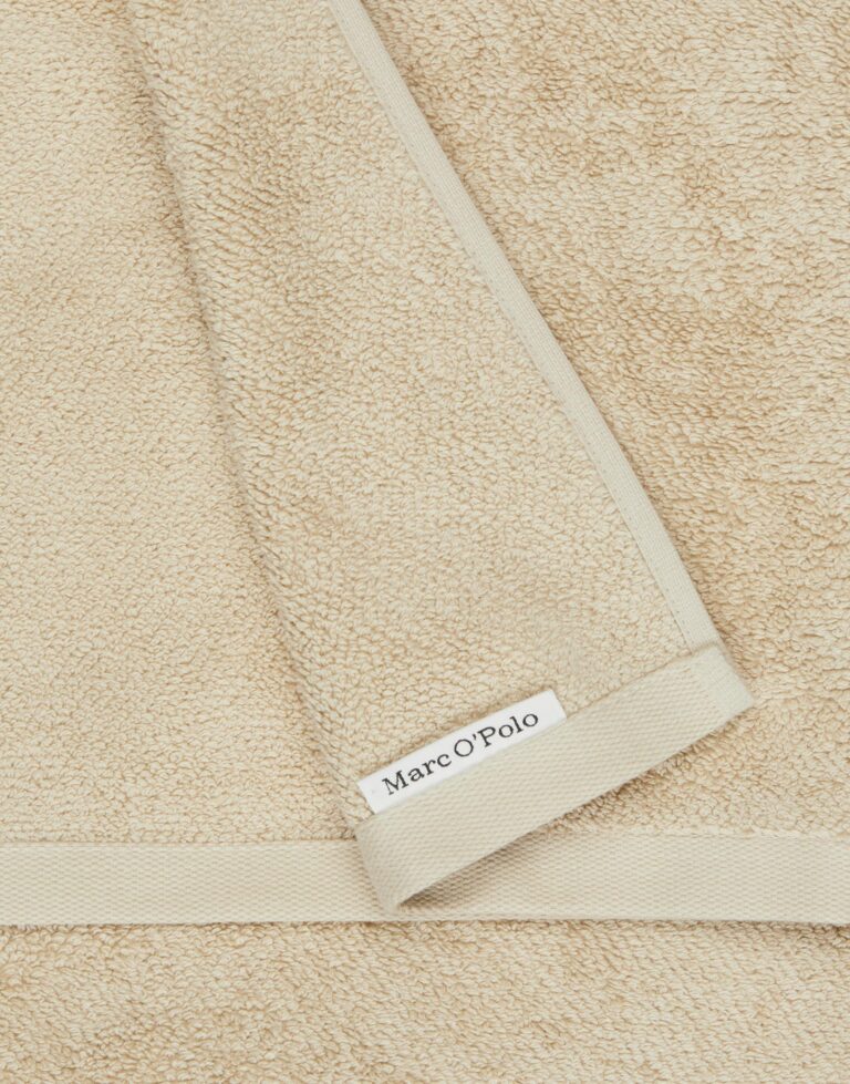 Het Marc O'Polo Timeless gastendoek in de kleur Dark Sand heeft een zachte beige structuur, nette randen en een label met het merklogo in de hoek.