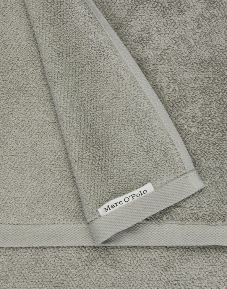 Een close-up van de Marc O'Polo Timeless gastendoek in grijs toont de lichtgrijze stof met structuur, een omgeslagen hoek en het label.