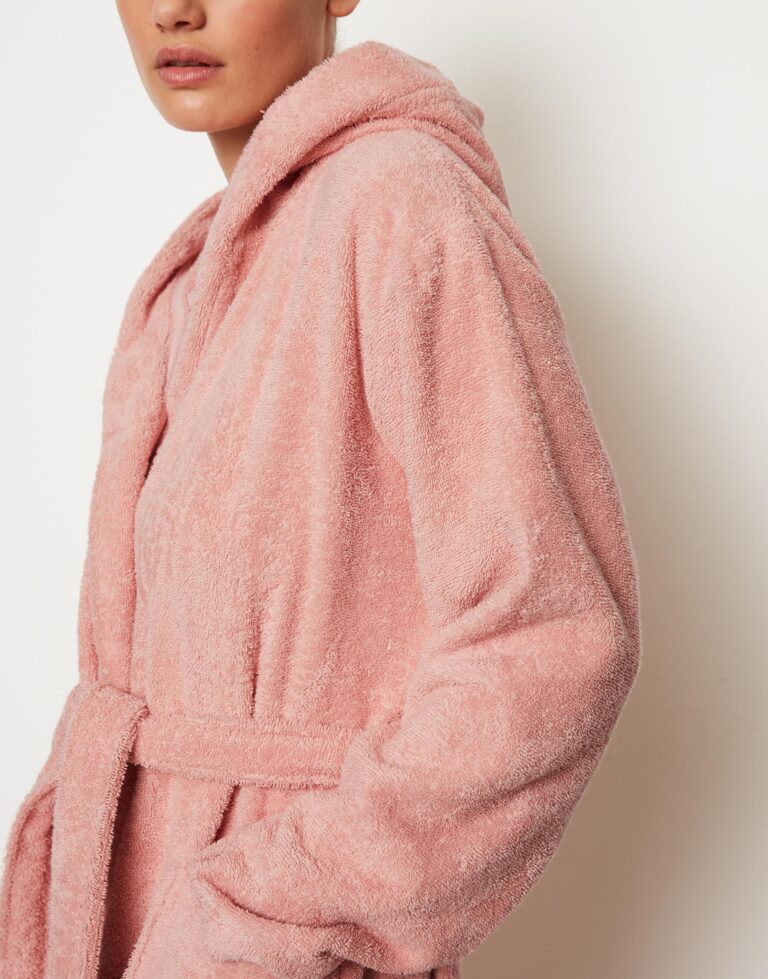 Een persoon draagt de Marc O'Polo Timeless Women-badjas in de kleur Rose Powder, gemaakt van zachte stof, met een capuchon en een ceintuur; het gezicht is gedeeltelijk zichtbaar.