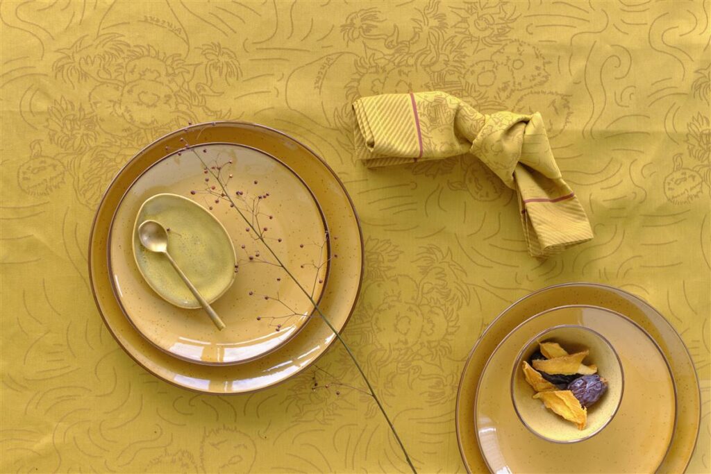 Een geel gedekte tafel met borden met patroon, een schaaltje gedroogd fruit, een gouden lepel en een servet op het Van Gogh Tournesol tafelkleed Yellow.