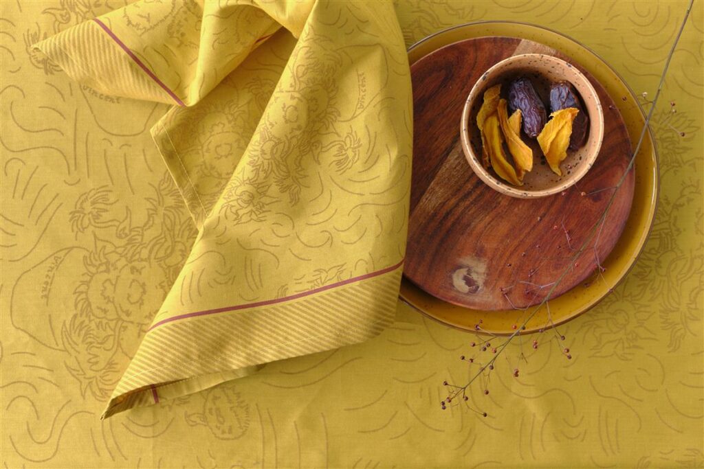 Het Van Gogh Tournesol-tafelkleed, geel met patroon en een roodgestreepte rand, ligt over de tafel met een houten schaal en rode bessen.