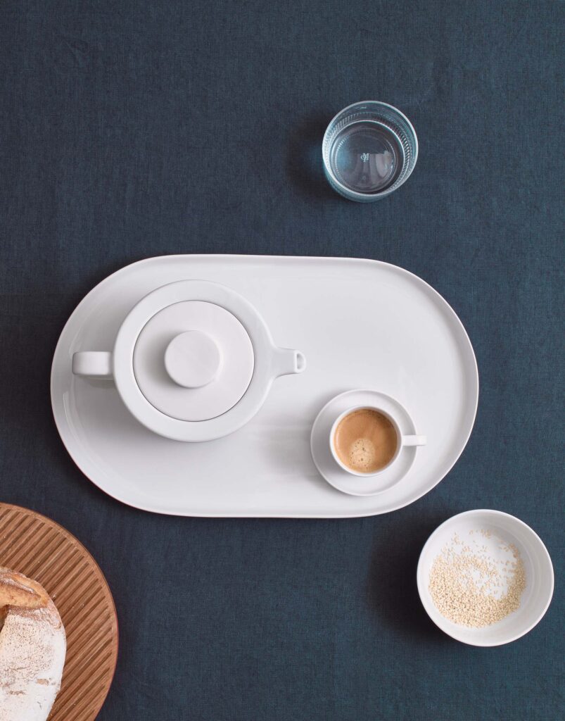Op de Marc O'Polo Valka tafelloper in indigoblauw staan een witte theepot, een espressokopje, sesamzaadjes en een glas water.