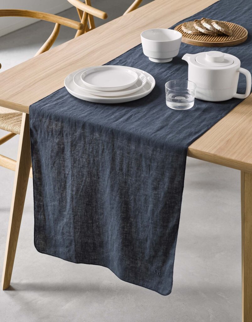 Een moderne eetkamer is gedekt met de Marc O'Polo Valka-tafelloper in indigoblauw, wit servies, een theepot en lichte houten stoelen.