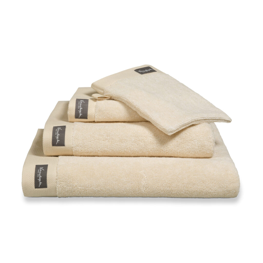 De Vandyck Home Mouline Badlaken Bleached Sand-handdoek is netjes opgestapeld en heeft een donker label met witte tekst.