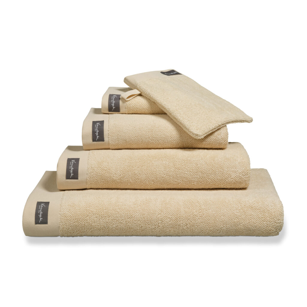 Het Vandyck Home Uni-badlaken in de kleur Bleached Sand is netjes opgevouwen en voorzien van een donker rechthoekig label met witte tekst.