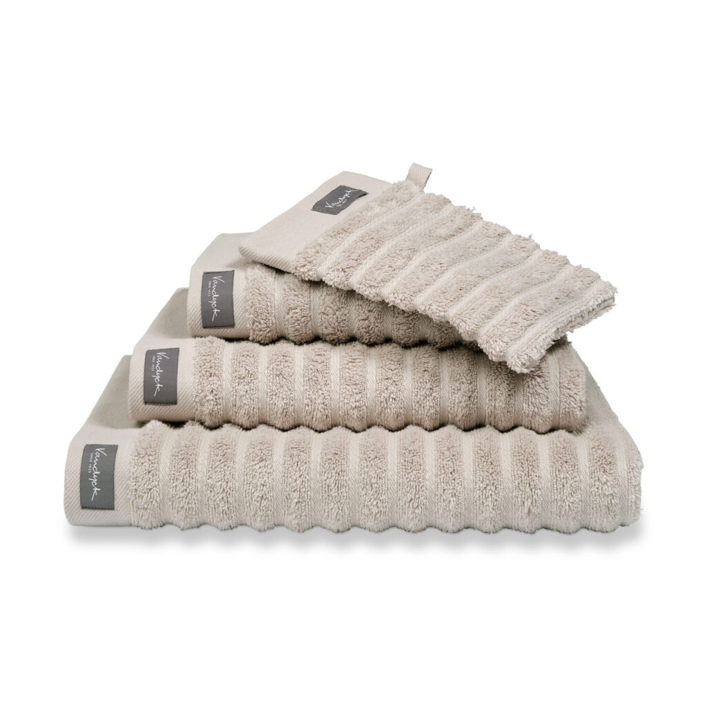 Een set Vandyck Home Wave-badhanddoeken in de kleur Stone, beige met ribbels en grijze labels, in verschillende maten van groot naar klein, inclusief washandje.