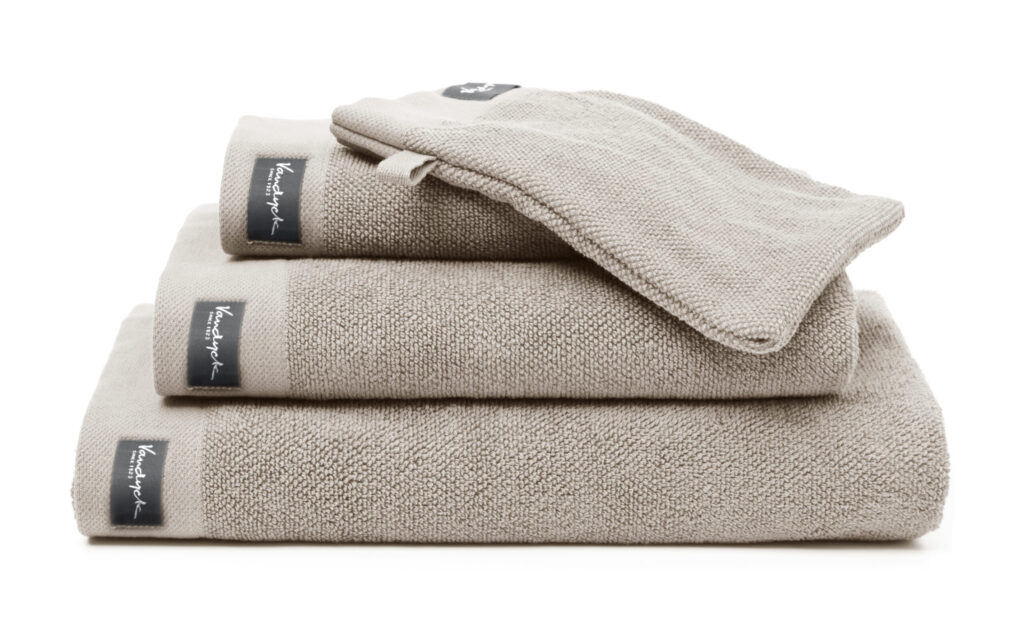 De Vandyck Home Uni Badlaken Stone-handdoek met zwart Kensington-label is netjes opgevouwen en wordt geleverd met een bijpassend washandje.