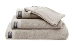 De Vandyck Home Uni Badlaken Stone-handdoek met zwart Kensington-label is netjes opgevouwen en wordt geleverd met een bijpassend washandje.