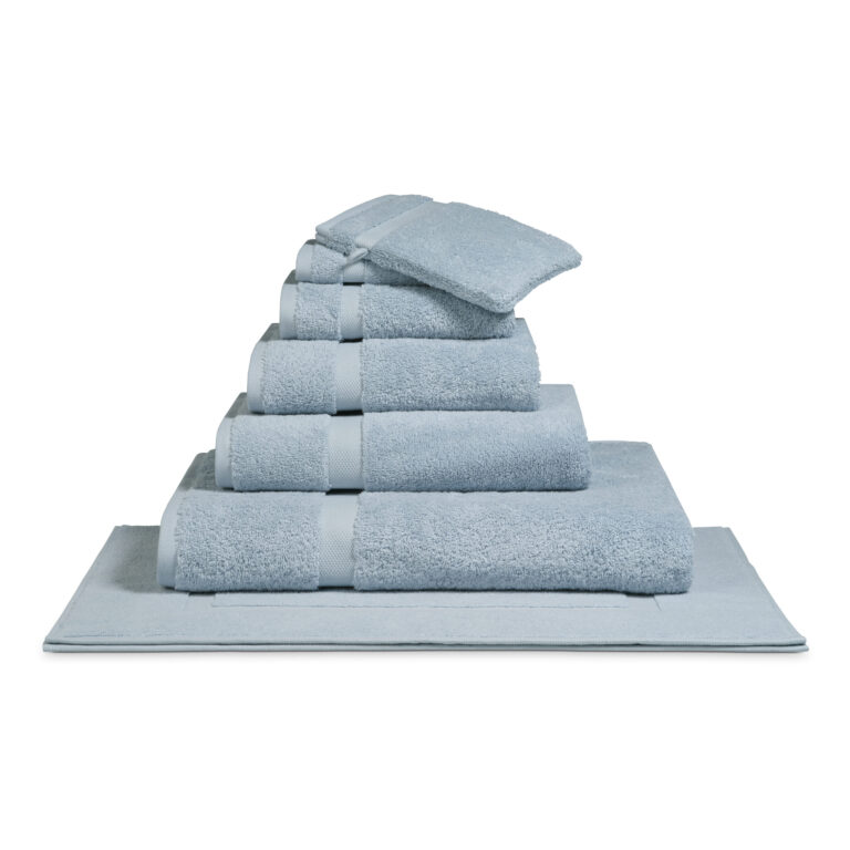 Het De Vandyck Ranger-badlaken in Misty Blue vormt samen met andere netjes opgevouwen handdoeken een piramide op een bijpassende badmat.