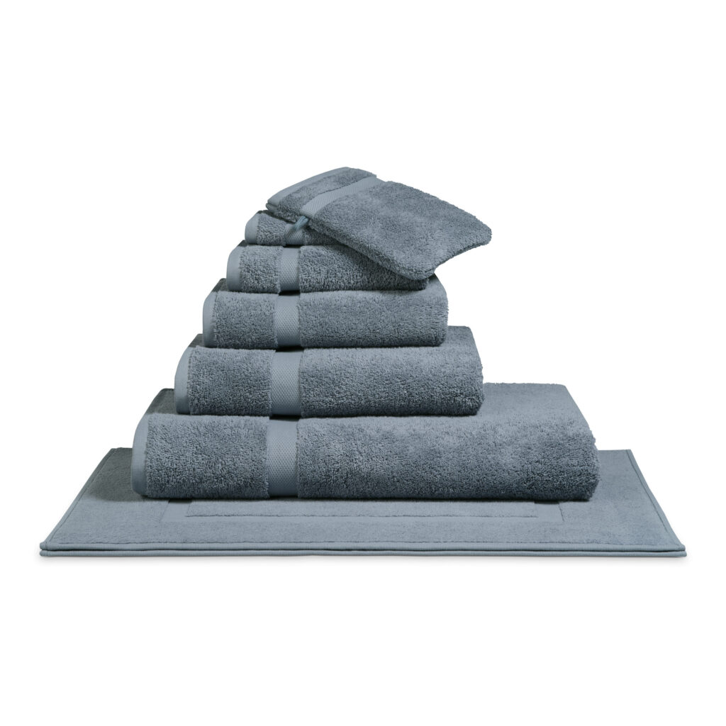 Het De Vandyck Ranger-badlaken in Dusty Blue ligt netjes opgestapeld, samen met bijpassende washandjes, op een badmat in blauwgrijze tinten.