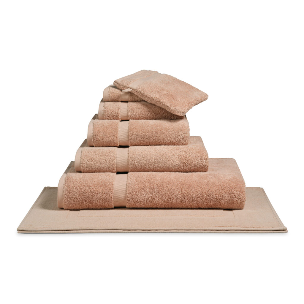 Op een beige badmat ligt het Vandyck Ranger-badlaken in de kleur Praline netjes opgestapeld, samen met bijpassende handdoeken en washandjes.