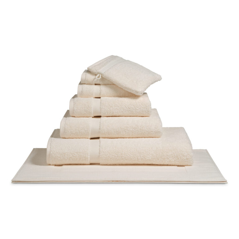 Het De Vandyck Ranger-badlaken in de kleur Cream tan ligt netjes opgestapeld in een piramidevorm met een lint eromheen, op een bijpassende badmat en washandje.
