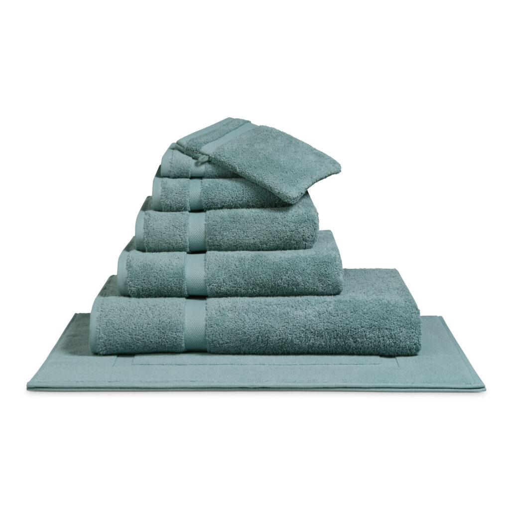 Het De Vandyck Ranger-badlaken in de kleur Mineral Blue ligt netjes opgevouwen op een bijpassende badmat en wordt stijlvol gepresenteerd.