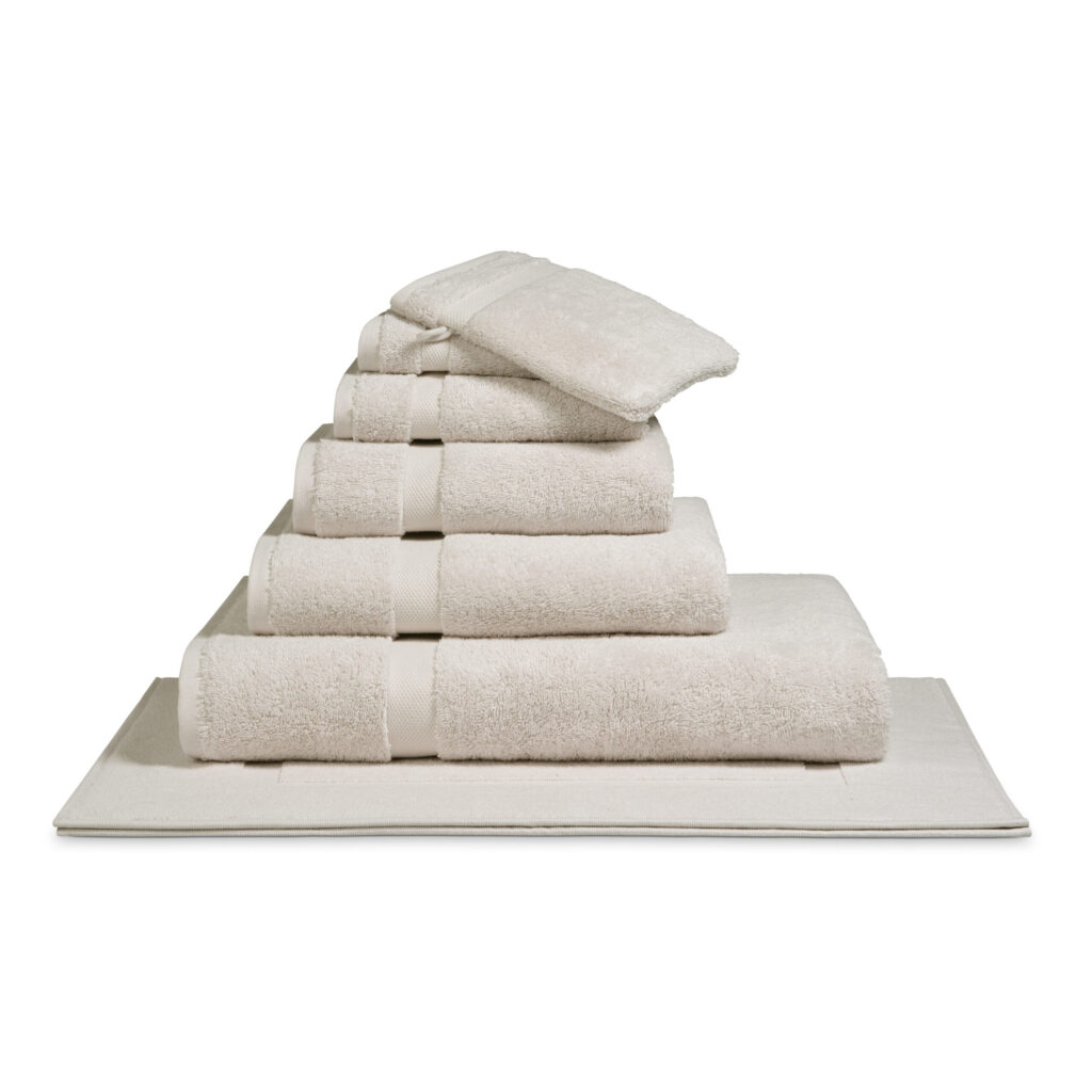 Het De Vandyck Ranger-badlaken in de kleur Cotton Cloud ligt netjes opgestapeld, samen met bijpassende beige handdoeken en washandjes, op een badmat.