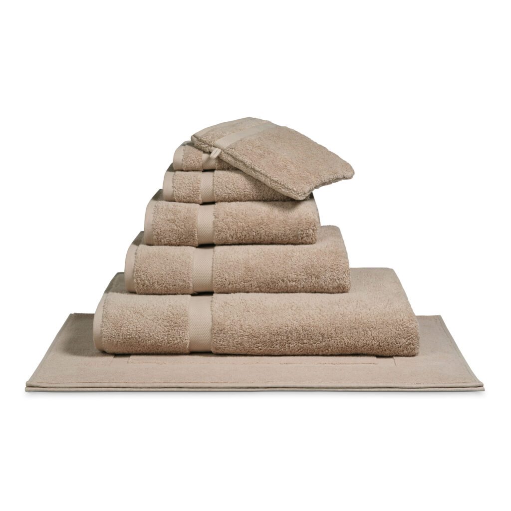 Het De Vandyck Ranger-badlaken in de kleur Almond ligt netjes opgestapeld met andere beige handdoeken in een piramidevorm op een bijpassende badmat.