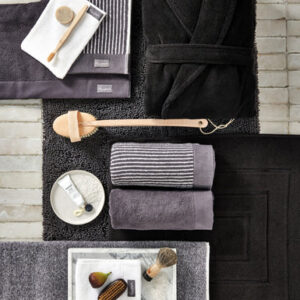 Op de zwarte en grijze badmatten liggen een Vandyck Home Mouline-badhanddoek in de kleur Off Black, een badjas en diverse verzorgingsproducten.
