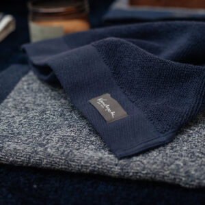 Het De Vandyck Home Mouline-badlaken in de kleur Blue Ink ligt netjes opgevouwen naast een grijze handdoek met het Leisure Club-label en een kaars erachter.