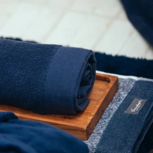 De Vandyck Home Mouline-handdoek Blue Ink ligt zorgvuldig opgerold op een houten dienblad, met ernaast een netjes opgevouwen exemplaar.