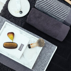 De Vandyck Home Mouline-handdoek in Off Black ligt netjes opgevouwen op een donkere ondergrond, met daarnaast luxe badkameraccessoires.