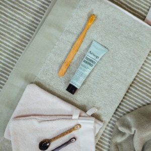 Op een opgevouwen Vandyck Home Mouline-handdoek in de kleur Smoke green liggen een bamboe-tandenborstel, Aesop-tandpasta, een beige washandje en drie lepels.