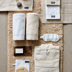 Op een beige mat liggen de Vandyck Home Petit Ligne-badhanddoeken in de kleur Bleached Sand, samen met handdoeken, zeep, een oogmasker en een badjas.