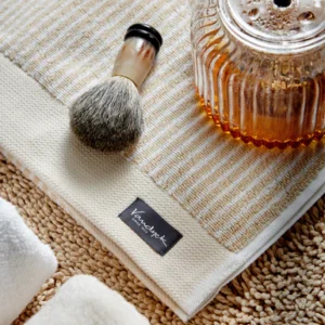 De Vandyck Home Petit Ligne Handdoek Bleached Sand ligt opgevouwen naast opgerolde handdoeken, een scheerkwast en een glazen flesje met bruine vloeistof op een gestructureerd oppervlak.