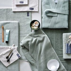 Spa-benodigdheden met de Vandyck Home Petit Ligne-handdoek in rookgroen, een bijpassende badjas, tandenborstel, tandpasta, maatlepels, schelpen in een schaal en een gestreept cadeauzakje.