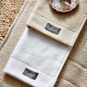 De Vandyck Home Uni-badhanddoek Bleached Sand en een witte handdoek met zwarte labels liggen netjes op een beige badmat naast badzout.