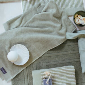 De Vandyck Home Uni Badlaken, een rookgroene handdoek, ligt netjes naast een beige handdoek, een gestreept washandje, een zeepje aan een touwtje, een schelpje met een bolletje en een schaaltje met sponsjes.