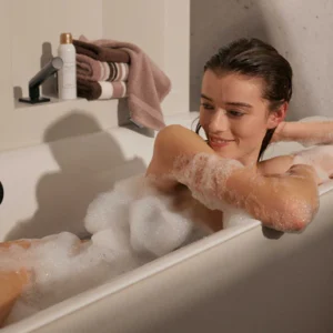 Een vrouw glimlacht terwijl ze ontspannen in een bubbelbad ligt en tegen de rand van het bad leunt. Een Vandyck Ranger-badlaken in de kleur Mauve ligt samen met een fles naast haar.