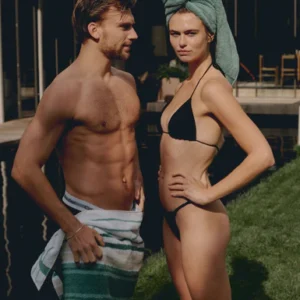 Een man met ontbloot bovenlijf en een handdoek om zijn middel staat buiten bij een zwembad en gebruikt het Vandyck Ranger-badlaken in de kleur Mineral Blue, terwijl naast hem een vrouw in een zwarte bikini staat met een groene handdoek op haar hoofd.