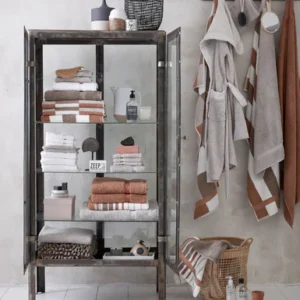 In een overzichtelijke badkamer met aardse tinten liggen Vandyck Ranger Handdoek Hazel-handdoeken mooi opgevouwen in een vitrinekast, omringd door zeepdispensers en decoratie. Naast de kast hangen badjassen aan haken en staat er een mand vol met extra handdoeken.