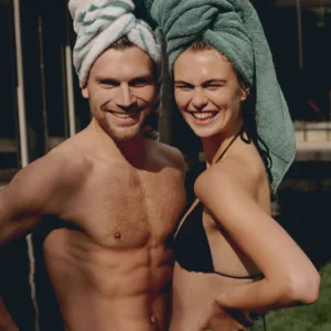 Een man en een vrouw in badkleding glimlachen ontspannen buiten naar de camera, met een Vandyck Ranger-handdoek in de kleur Mineral Blue om hun hoofd.