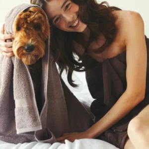 Een lachende vrouw zit gezellig op het bed met haar bruine hond; ze zijn allebei knus gewikkeld in de Vandyck Ranger Handdoek Rose taupe.