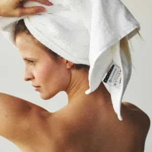 Een vrouw met blote schouders staat in profiel afgebeeld terwijl ze haar natte haar in een Vandyck Ranger Handdoek White wikkelt, waarvan het label zichtbaar is.