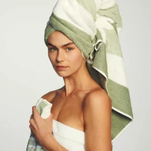 Een persoon wikkelt de Vandyck Vancouver-handdoek in de kleur Smoke Green om haar haar en lichaam en kijkt recht in de camera.