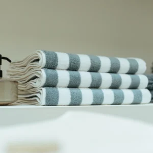 De Vandyck Verona Stripes Handdoek: parelblauwe handdoeken met witte en blauwe strepen liggen netjes opgestapeld naast een zeepdispenser.