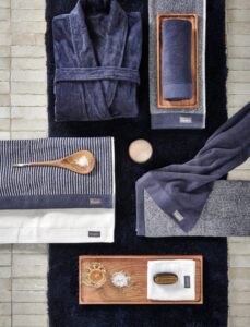 De Vandyck Toronto badmat Blue Ink ligt op de vloer, in combinatie met marineblauwe en witte handdoeken, een badjas en spa-accessoires.