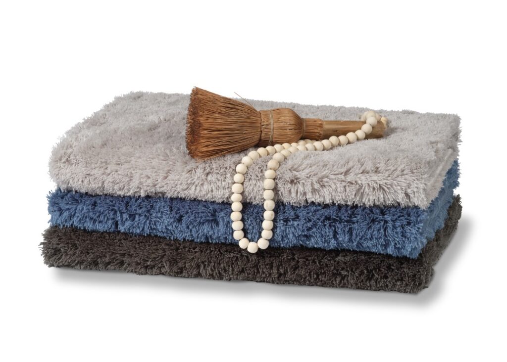 De Vandyck Toronto Badmat Vintage Blue bestaat uit drie opgevouwen, zachte handdoeken in grijs, blauw en donkergrijs, met daarop een klein strooien borsteltje en witte kralen.