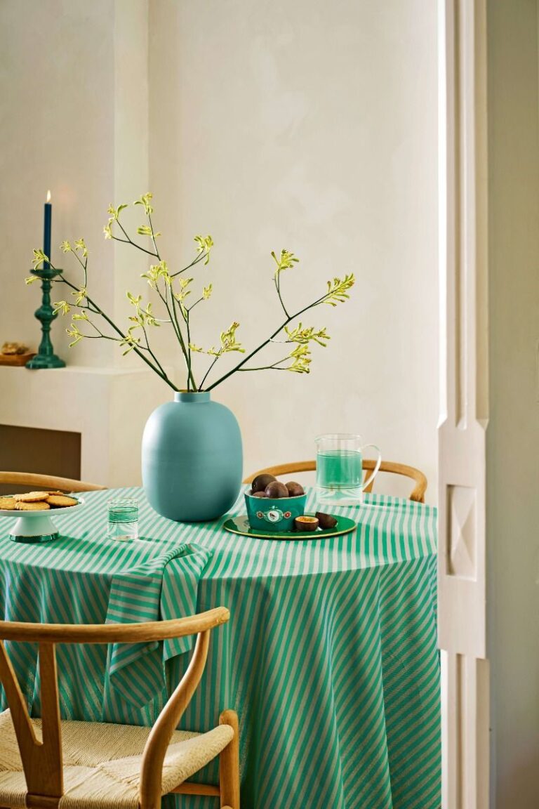 Op de rondgedekte tafel met een groen-wit gestreept tafelkleed staan een blauwe vaas met gele bloemen, servies van Pip Studio en een brandende kaars voor een gezellige sfeer.