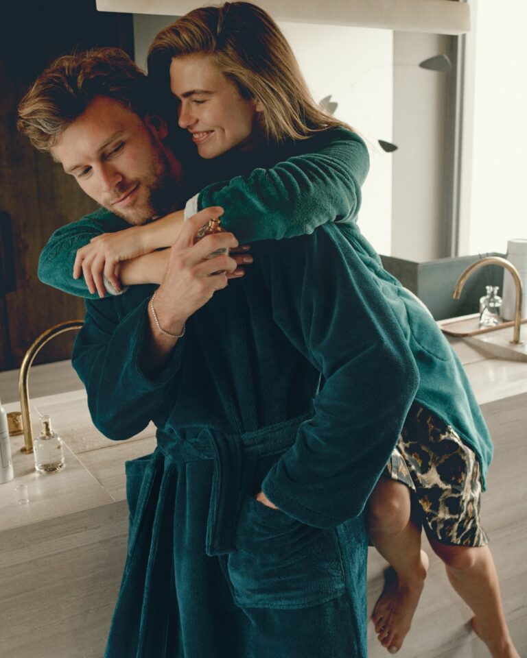 In een gezellige, moderne badkamer knuffelt een glimlachende vrouw in een groene badjas speels een man in een Vandyck Beaumont-badjas in de kleur ‘dark sea’, terwijl hij bij de wastafel staat met een spuitfles.
