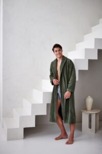 Iemand draagt een Vandyck Beaumont-badjas in saliegroen met een korte broek en staat op blote voeten bij moderne traptreden, naast een tafeltje met een vaas.