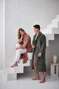 Een vrouw in een roze badjas met een witte broek zit op de trap; naast haar staat een man in de Vandyck Beaumont-badjas in de kleur sage green, maat S. Op een lagere trede staat een witte vaas.